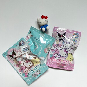 May include: Two small plastic bags of Hello Kitty erasers. The bags are blue and pink with Hello Kitty and her friends on them. The text on the bags reads "Hello Kitty Erasers" in English and Chinese. A small Hello Kitty eraser is sitting on top of the bags.