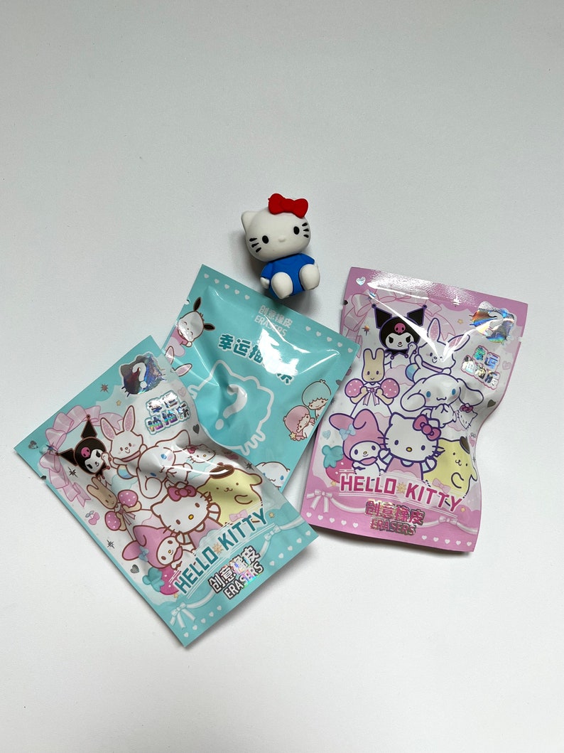 May include: Two small plastic bags of Hello Kitty erasers. The bags are blue and pink with Hello Kitty and her friends on them. The text on the bags reads "Hello Kitty Erasers" in English and Chinese. A small Hello Kitty eraser is sitting on top of the bags.