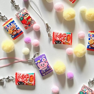 Op de afbeelding: Een verzameling kleurrijke, miniatuur snoepreepcharms met sleutelhangers. De charms tonen populaire Japanse snacks zoals KitKat, Pocky en een aardbeienkoekje. De charms zijn bevestigd aan roze, witte en gele pompoenen.
