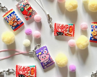 Puede incluir: Una colección de encantos de barras de chocolate en miniatura de colores con llaveros. Los encantos presentan aperitivos japoneses populares como KitKat, Pocky y una galleta con sabor a fresa. Los encantos están unidos a pompones rosas, blancos y amarillos.