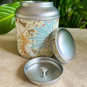 Peut inclure: Une boîte de rangement cylindrique en métal avec une finition argentée et un couvercle amovible. L'extérieur présente un motif botanique répétitif dans des tons de bleu sarcelle, jaune moutarde et blanc. Un couvercle séparé avec un bouton est inclus.