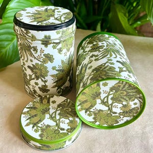Peut inclure: Deux boîtes cylindriques en métal et un couvercle circulaire séparé présentant un fond blanc avec un motif floral détaillé en vert olive et vert foncé. Les boîtes ont une finition lisse et un bord vert, avec un couvercle posé dessus.