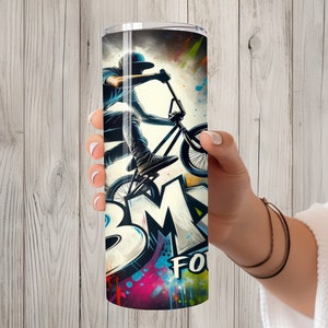 May include: A stainless steel tumbler with a colorful graphic of a person riding a BMX bike. The graphic includes the word "BMX" in large, stylized letters. The background is a mix of colorful paint splatters.