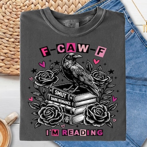 Puede incluir: Camiseta gris oscuro con un diseño de cuervo sobre libros con las palabras "Smut" y "Dark Romance". La camiseta también presenta rosas, corazones, estrellas y los textos "F-CAW-F" y "I'M READING".