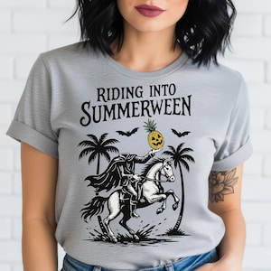 May include: A light gray t-shirt with the words "RIDING INTO SUMMERWEEN" above a black and white illustration. The illustration depicts a headless figure on a horse holding a jack-o'-lantern pineapple, with palm trees and bats.