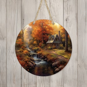 May include: A round wooden wall hanging featuring a painting of a small cottage nestled in a woodland scene. The cottage is surrounded by trees with orange, yellow, and red leaves. A stream flows in front of the cottage with a waterfall. Sunlight streams through the trees.