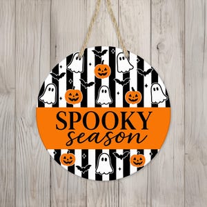 May include: Round Halloween sign with a black and white striped background, featuring orange pumpkins, white ghosts, and black bats. The sign has the words "Spooky Season" in orange and black.