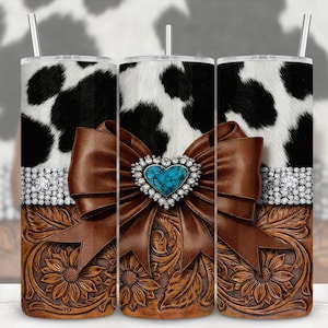 May include: A stainless steel tumbler with a cowhide pattern, a brown leather bow with a turquoise heart-shaped jewel, and a floral embossed leather base. The tumbler has a silver straw and a rhinestone band.