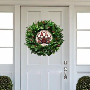 May include: A Christmas wreath with a circular gnome design at the center. The wreath is made of green foliage, red berries, and a round sign featuring two gnomes. The door is white with a silver handle.