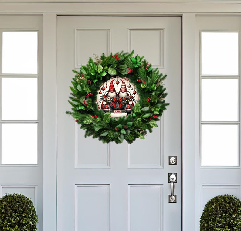 May include: A Christmas wreath with a circular gnome design at the center. The wreath is made of green foliage, red berries, and a round sign featuring two gnomes. The door is white with a silver handle.
