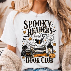 May include: A white t-shirt featuring a graphic print with the text SPOOKY READERS BOOK CLUB and running on boos, brews & books. The design includes a ghost reading a book, a stack of books, a black cat, a mug, a lantern, and a pumpkin.