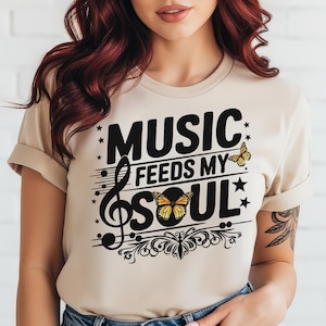 May include: Beige t-shirt with the text "MUSIC FEEDS MY SOUL" in black, featuring a treble clef, butterflies, and stars. The shirt has rolled-up sleeves and is made of a soft material.