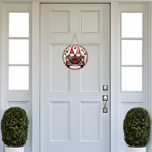 May include: A round wooden door sign featuring a festive gnome design. The sign has a red and white border and a jute rope for hanging. The image shows two gnomes with red hats and beards, surrounded by Christmas decorations.