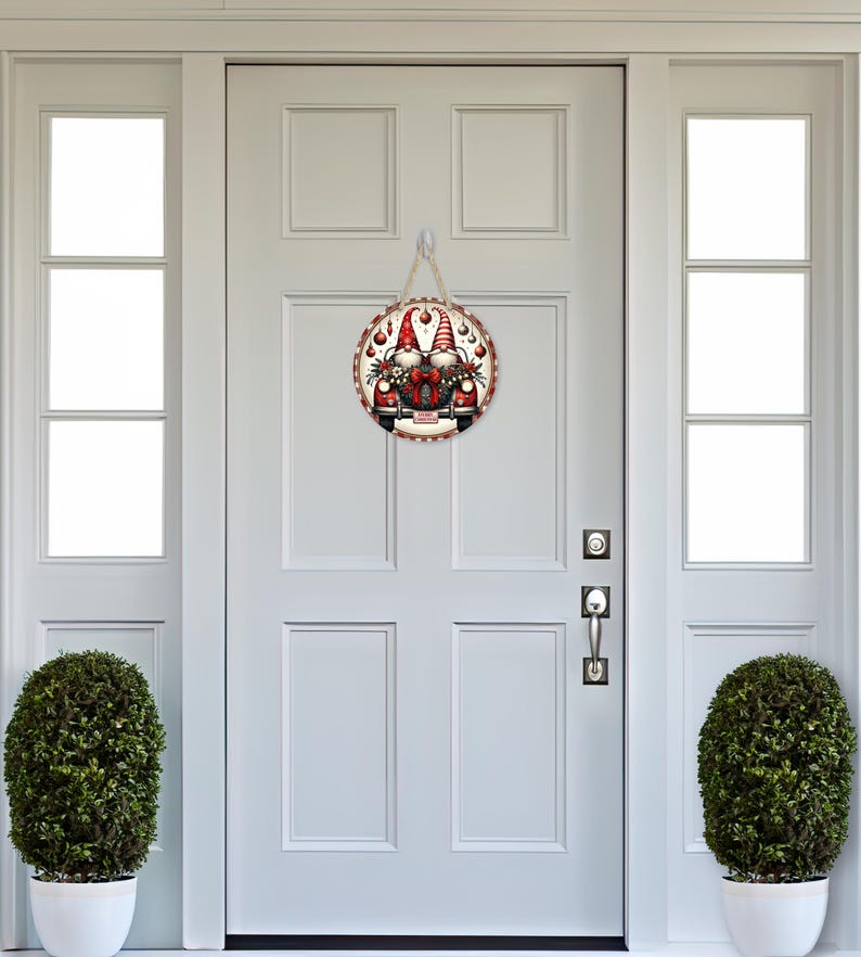 May include: A round wooden door sign featuring a festive gnome design. The sign has a red and white border and a jute rope for hanging. The image shows two gnomes with red hats and beards, surrounded by Christmas decorations.