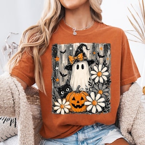 May include: A burnt orange t-shirt featuring a graphic print of a white ghost wearing a black witch hat with an orange bow, a black cat, an orange jack-o'-lantern, and white daisies against a background of black and white vertical stripes.
