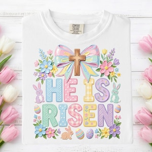 May include: White t-shirt with a colorful Easter design. The design includes a cross, bow, flowers, bunnies, and the words "HE IS RISEN" in pastel colors. A Comfort Colors tag is visible.