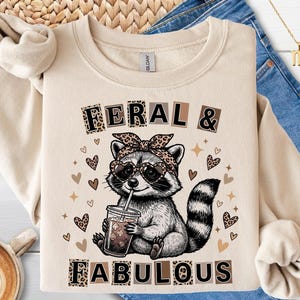Peut inclure: Sweat-shirt beige avec un dessin de raton laveur portant des lunettes de soleil en forme de cœur et un nœud léopard. Le texte "FERAL & FABULOUS" est au-dessus et en dessous du raton laveur. Le design comprend des cœurs et des étoiles.