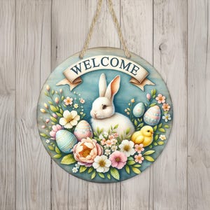 May include: Round wooden Easter sign with a white rabbit, colorful eggs, and a yellow chick surrounded by flowers. The sign has a banner that reads "WELCOME" and is hanging by a jute rope. The background is a light blue circle with a wooden texture.