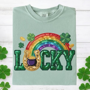 以下が含まれることがあります： 聖パトリックの日のデザインが施された、明るい緑色のTシャツ。虹、金の入った蹄鉄、クローバー、緑のスパンコールで「LUCKY」の文字が描かれています。Tシャツはクルーネックです。