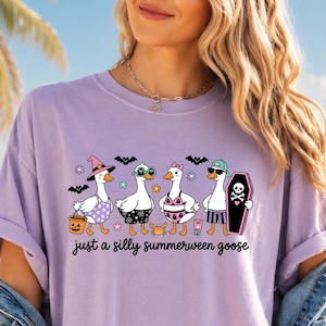 Op de afbeelding: Lichtpaars T-shirt met een leuke afbeelding van vier ganzen in Halloween- en zomeroutfits. De ganzen dragen zonnebrillen, hoeden en badkleding. De tekst "just a silly summerween goose" staat eronder.
