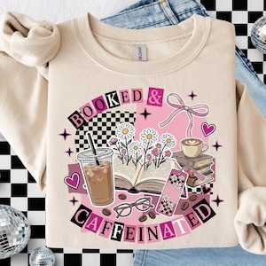 May include: Beige sweatshirt with a graphic design featuring the words "BOOKED & CAFFEINATED" in a pink and black checkerboard font. The design includes coffee, books, flowers, and hearts.