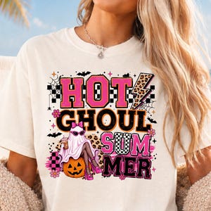 Op de afbeelding: Een crèmekleurig t-shirt met een grafische print met de tekst "HOT GHOUL SUMMER". Het ontwerp bevat een geest met een roze zonnebril en een strik, zittend op een pompoen, omringd door luipaardprint, dambordpatronen en vleermuizen.