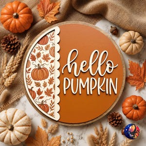 May include: A round wooden sign with a burnt orange background and the white text hello PUMPKIN. The left side features a scalloped edge panel with a pattern of orange pumpkins and brown autumn leaves on a cream background.