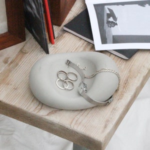 Op de afbeelding: Een grijze betonnen schaal met een afgeronde vorm, ontworpen om sieraden in te bewaren. De schaal bevat een zilveren armband, een zilveren ketting en vier zilveren ringen.