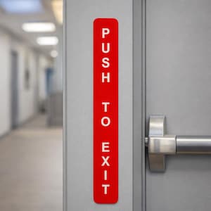 May include: A red rectangular sign with the words "PUSH TO EXIT" in white capital letters is affixed to a gray door. The door has a silver handle. The background shows a hallway with a blurred perspective.