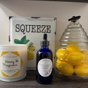 K&ouml;nnte beinhalten: Eine blaue Glasflasche mit Tropfer und einem wei&szlig;en Etikett, auf dem "Little Rock Creek Co. Palma Christi Rosehip Serum Est. 2021" steht. Die Flasche steht auf einer Holzoberfl&auml;che neben einer wei&szlig;en Kerze mit einem schwarzen und goldenen Etikett, auf dem "Red Leaf Home Honey Magnolia Coconut and Beeswax Blend with Essential Oils" steht. Ein Glasgef&auml;&szlig; mit einem Bienenstockmuster ist mit gelben, zitronenf&ouml;rmigen Badebomben gef&uuml;llt.