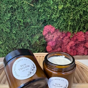May include: Two amber glass jars with white labels that read "Little Rock Creek Co. Sun Tallow Est. 2025". The jars are filled with a white cream and are sitting on a woven wicker surface.