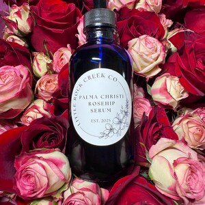 K&ouml;nnte beinhalten: Eine blaue Glasflasche mit Tropfer, umgeben von roten und wei&szlig;en Rosen. Das Flaschenetikett lautet "Little Rock Creek Co. Palma Christi Rosehip Serum Est. 2025".