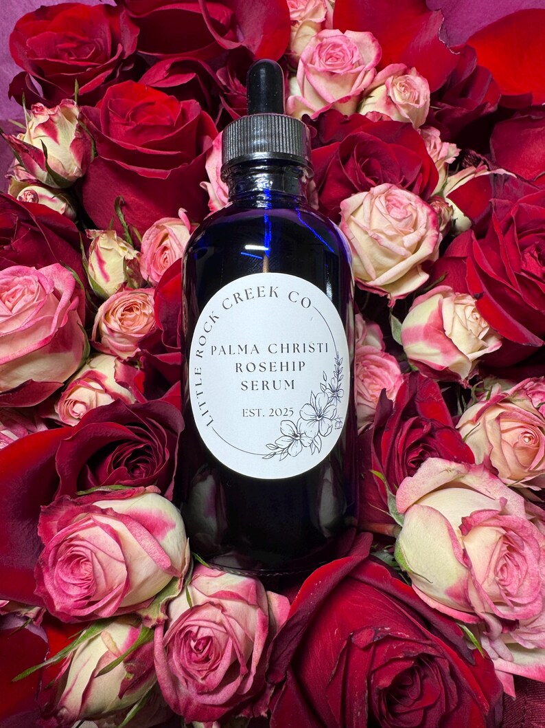 K&ouml;nnte beinhalten: Eine blaue Glasflasche mit Tropfer, umgeben von roten und wei&szlig;en Rosen. Das Flaschenetikett lautet "Little Rock Creek Co. Palma Christi Rosehip Serum Est. 2025".