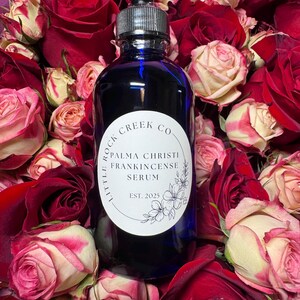 K&ouml;nnte beinhalten: Eine blaue Glasflasche mit Tropfer und einem wei&szlig;en Etikett mit der Aufschrift "Little Rock Creek Co. Palma Christi Frankincense Serum Est. 2025". Die Flasche ist von roten und wei&szlig;en Rosen umgeben.