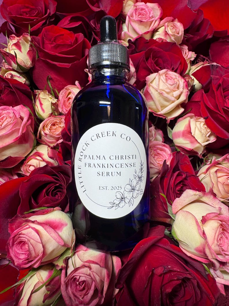 K&ouml;nnte beinhalten: Eine blaue Glasflasche mit Tropfer und einem wei&szlig;en Etikett mit der Aufschrift "Little Rock Creek Co. Palma Christi Frankincense Serum Est. 2025". Die Flasche ist von roten und wei&szlig;en Rosen umgeben.