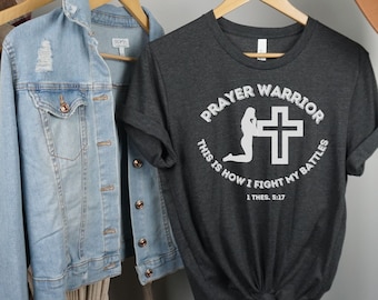 May include: A dark gray t-shirt with a white graphic design that says "Prayer Warrior" and "This is how I fight my battles 1 Thess. 5:17". The graphic features a silhouette of a person kneeling in prayer with a cross behind them.