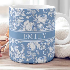 Puede incluir: Taza de cerámica blanca con un estampado floral azul y el nombre "EMILY" en letras blancas. La taza tiene un asa blanca y una banda azul claro. El diseño floral incluye rosas y hojas blancas.