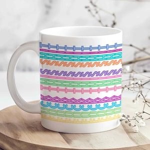 May include: White ceramic mug with a colorful geometric pattern. The design features horizontal bands of various shapes and colors, including blue, orange, green, pink, and purple. The mug has a curved handle and sits on a wooden surface.