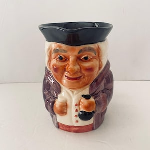 May include: A ceramic character jug shaped like a person wearing a black tricorn hat, a purple coat, and a white vest. The figure has a tan complexion, rosy cheeks, and is holding a small black object in one hand. The jug features a glossy finish.