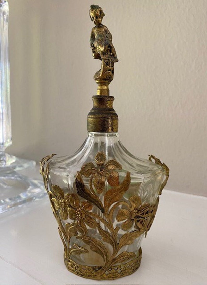 May include: A vintage glass perfume bottle with a gold-toned metal floral design and a cherub-shaped stopper.