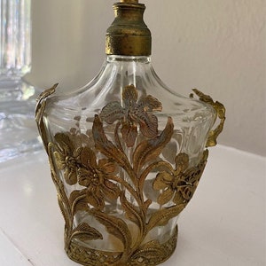 May include: A clear glass perfume bottle with a gold metal floral design. The bottle has a gold metal stopper and a gold metal base.