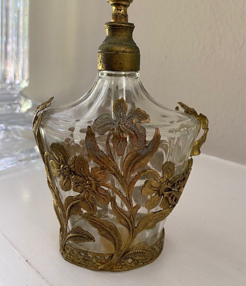 May include: A clear glass perfume bottle with a gold metal floral design. The bottle has a gold metal stopper and a gold metal base.