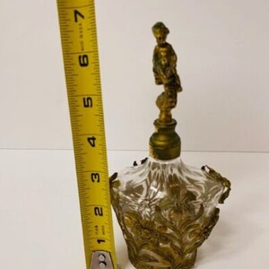 May include: A clear glass perfume bottle with a gold-toned metal top and a decorative floral design. The bottle stopper features a small gold-toned figurine of a woman.