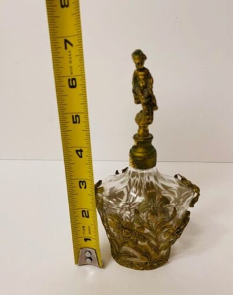 May include: A clear glass perfume bottle with a gold-toned metal top and a decorative floral design. The bottle stopper features a small gold-toned figurine of a woman.