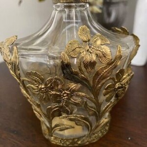 May include: A clear glass bottle with a gold top and intricate gold floral designs. The bottle is empty and has a vintage look.