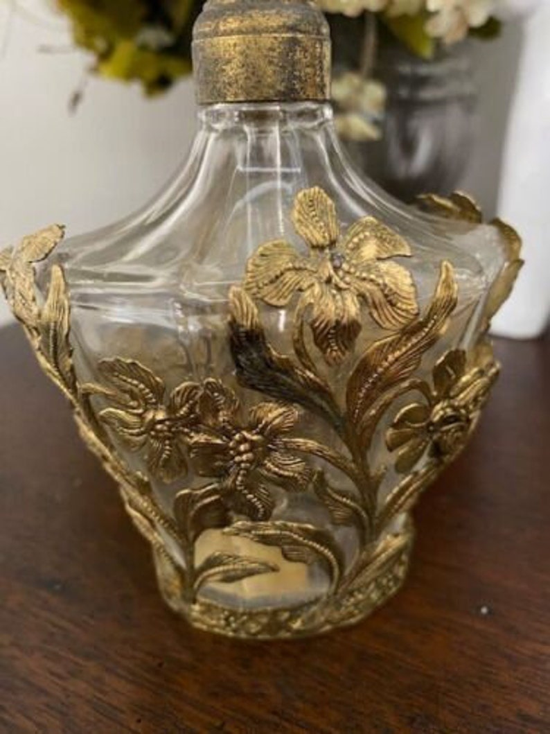 May include: A clear glass bottle with a gold top and intricate gold floral designs. The bottle is empty and has a vintage look.