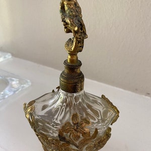 May include: A vintage glass perfume bottle with a gold-toned cherub figure on top. The bottle is decorated with gold-toned floral designs.