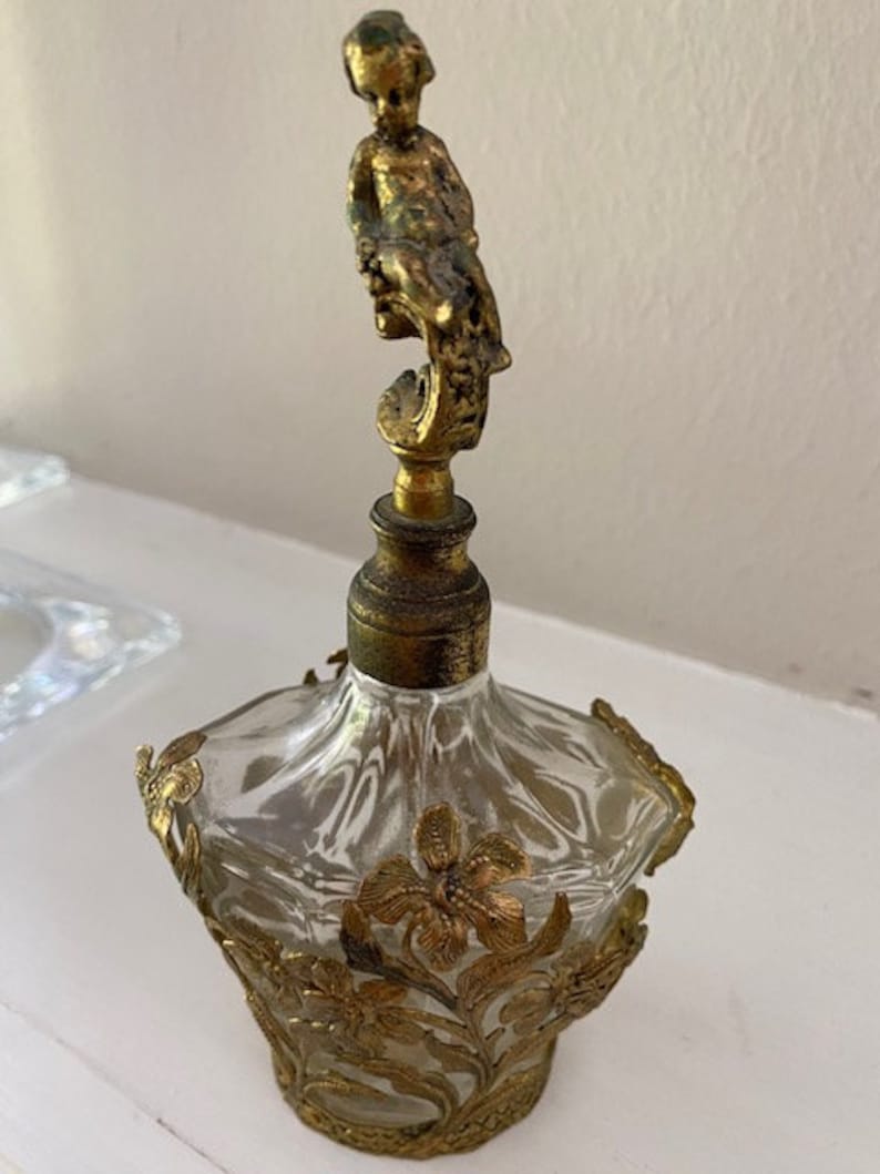 May include: A vintage glass perfume bottle with a gold-toned cherub figure on top. The bottle is decorated with gold-toned floral designs.