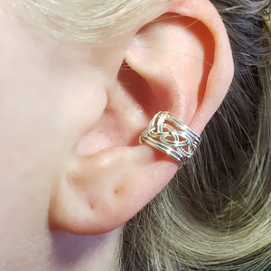 May include: Silver ear cuff with a woven knot design. The cuff is worn on the upper part of the ear.