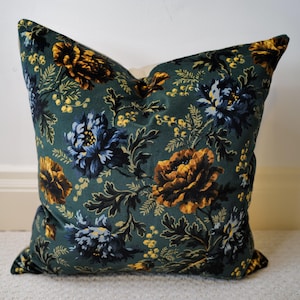 May include: Square decorative pillow with a dark teal background and a floral pattern. The flowers are in shades of gold, navy blue, and cream. The pillow is made of a soft fabric and is likely used for home decor.